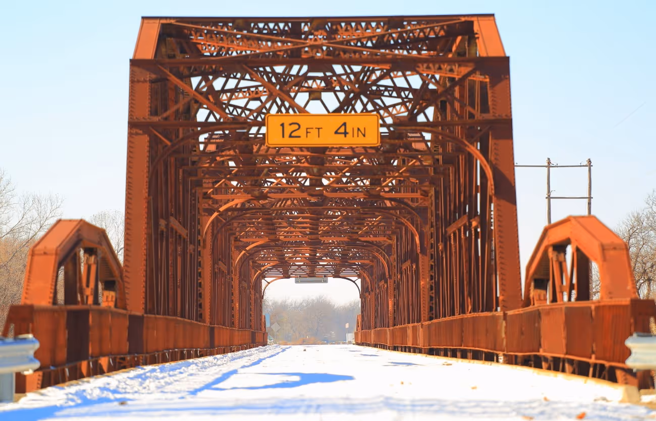 Rust-colored metal truss bridge with a snow-covered road and a height clearance sign of 12 feet 4 inches.