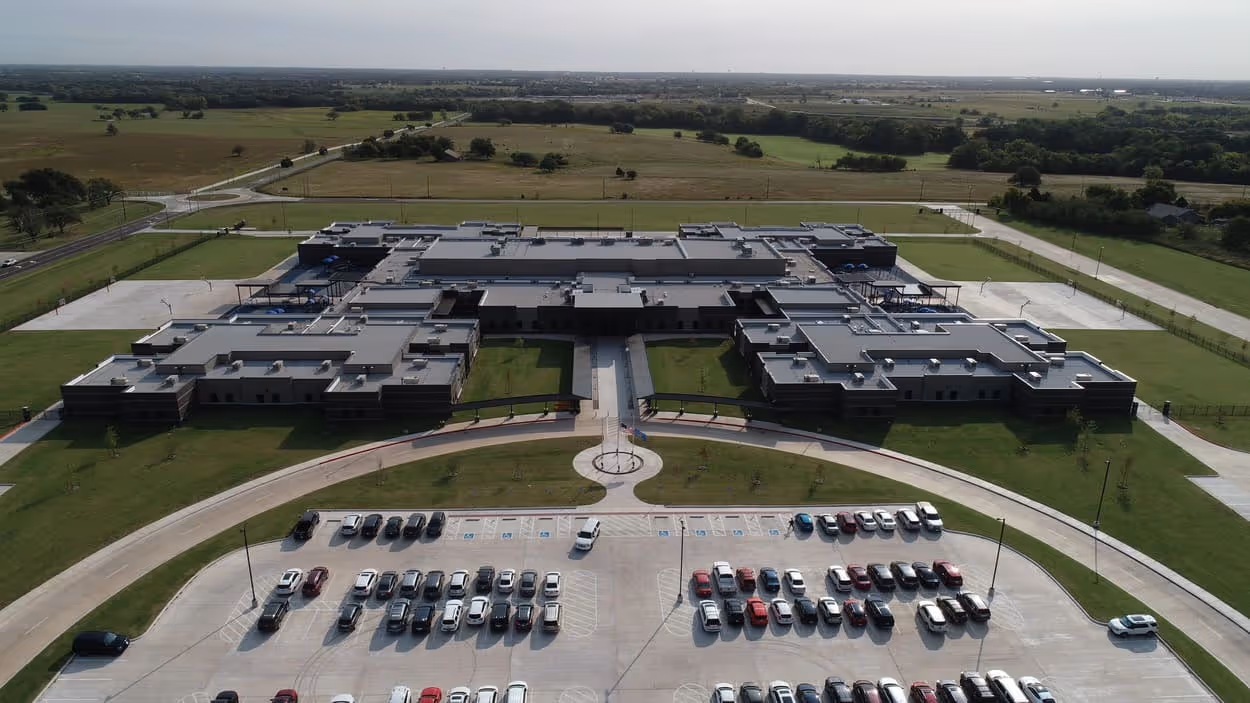 Aerial view of a large, single-story school building with multiple wings, surrounded by green fields and parking lots filled with cars.