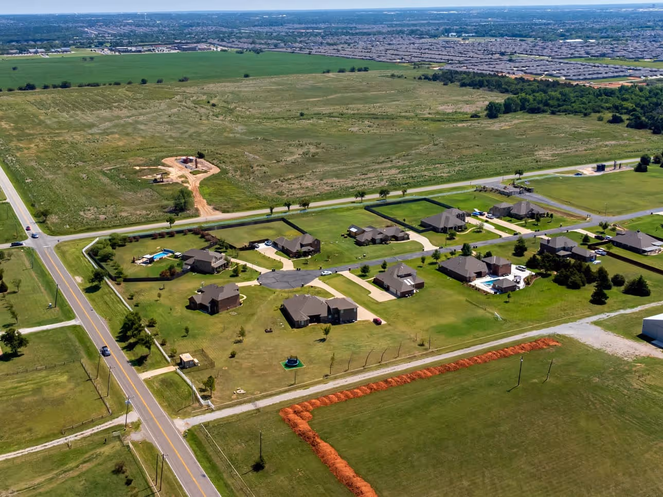 Aerial view of a suburban neighborhood with spacious single-family homes, green lawns, and intersecting roads surrounded by open fields.