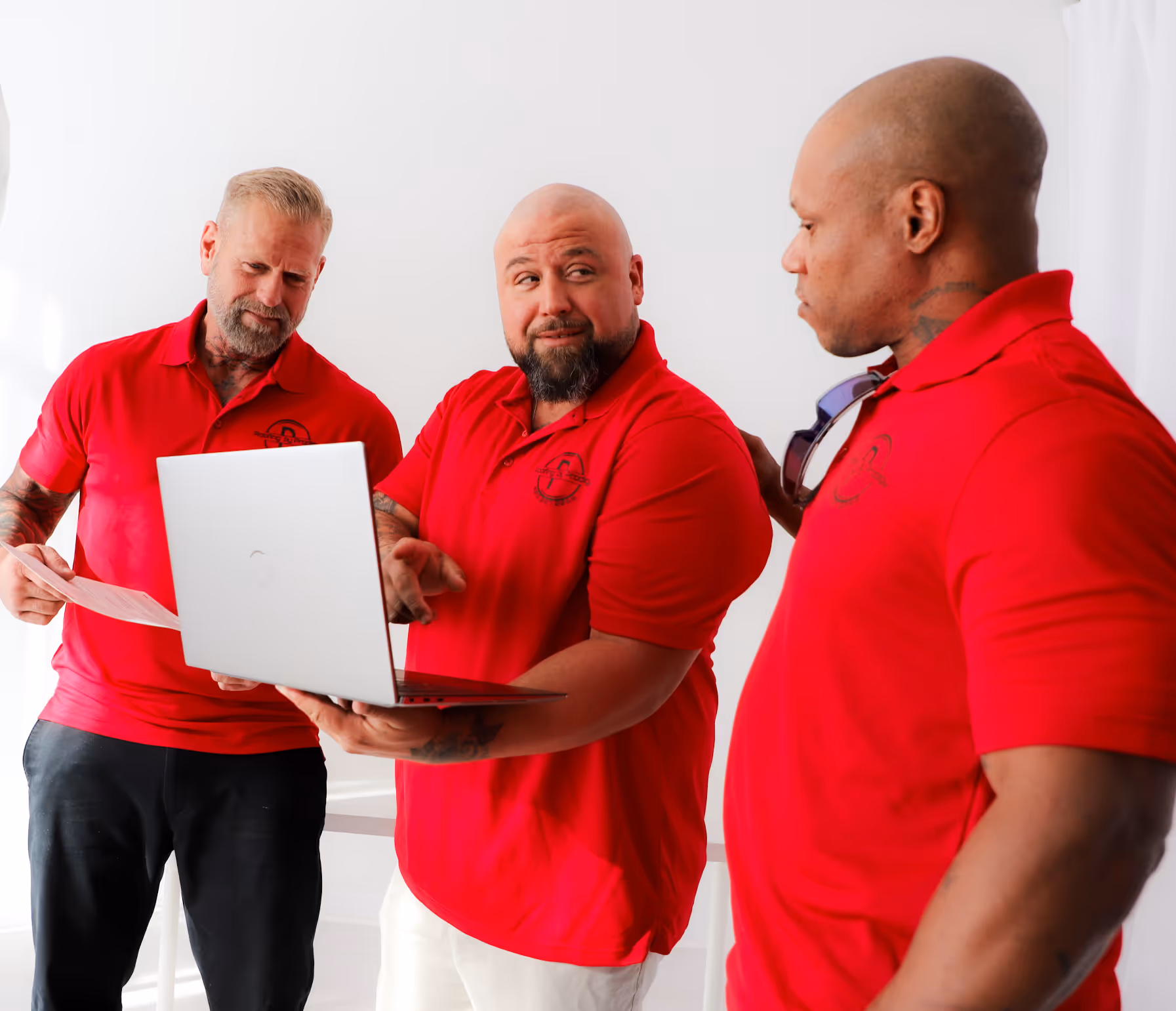 Roofing by Prodigy - Two men in red shirts laughing together at a white desk with a laptop, plant, cap, and cup.