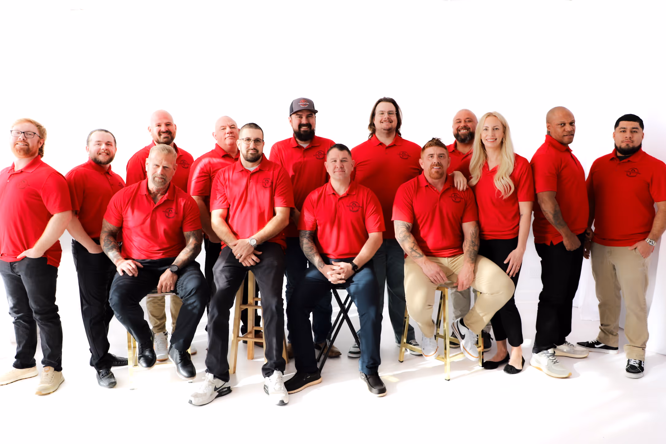 Roofing by Prodigy - Group of fourteen people wearing matching red polo shirts posing on stools and standing against a white background.