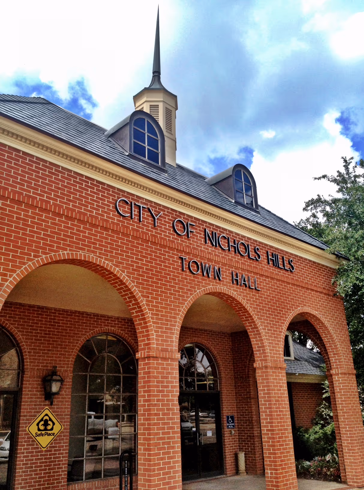 Brick building entrance with arched doorways and sign reading City of Nichols Hills Town Hall under a sky with scattered clouds.