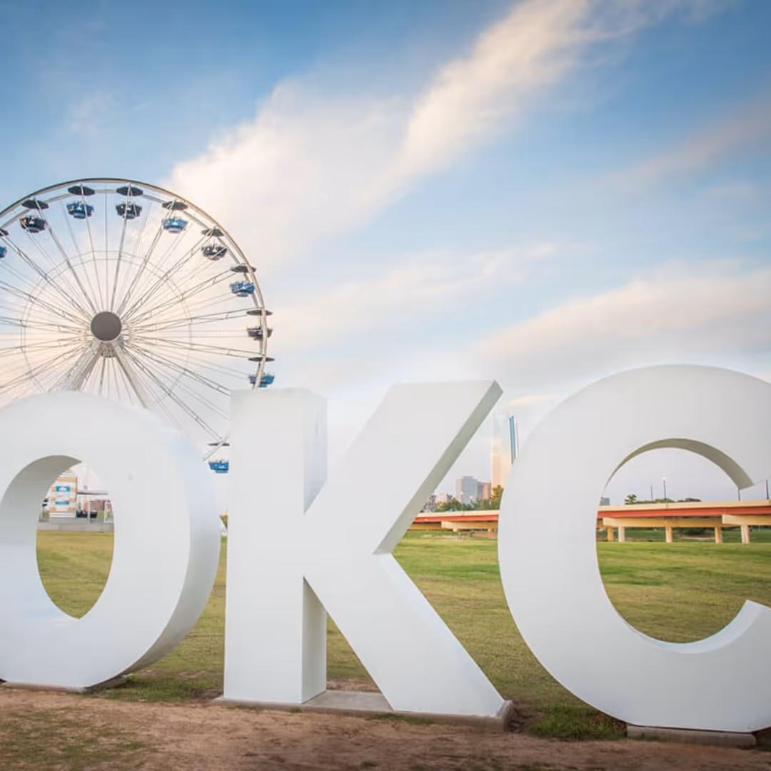 Large white OKC letters sculpture on grass with a Ferris wheel and blue sky in the background.