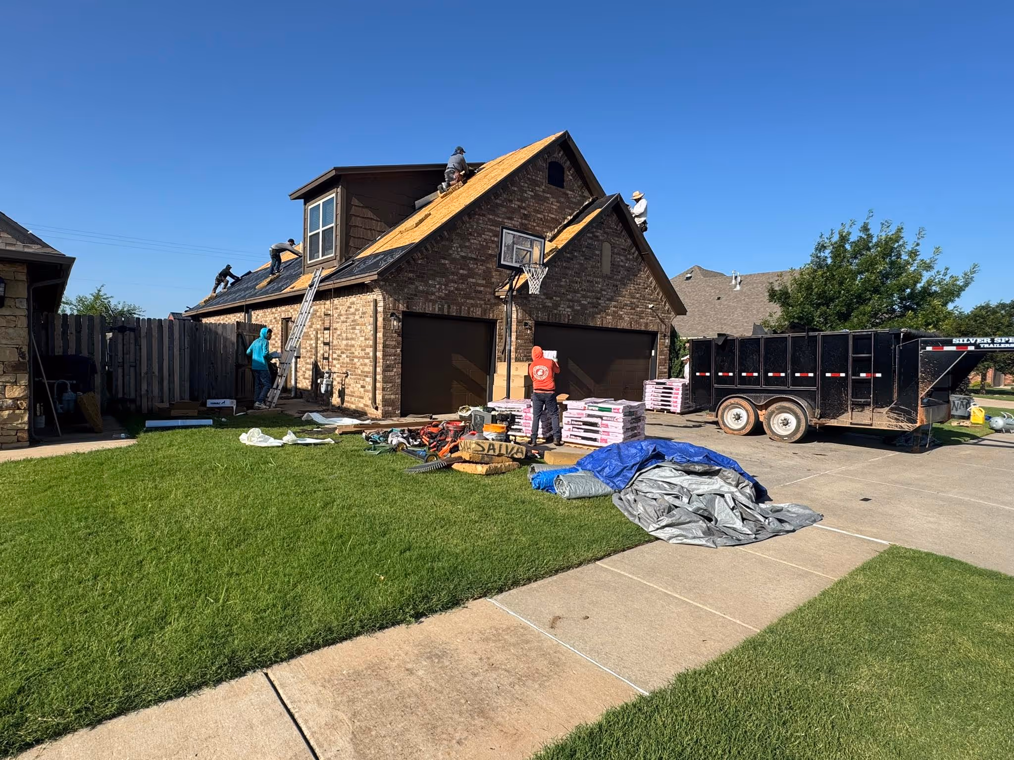 Roofing by Prodigy - Several workers installing a new roof on a two-car garage with roofing materials stacked nearby and a black trailer parked in the driveway.