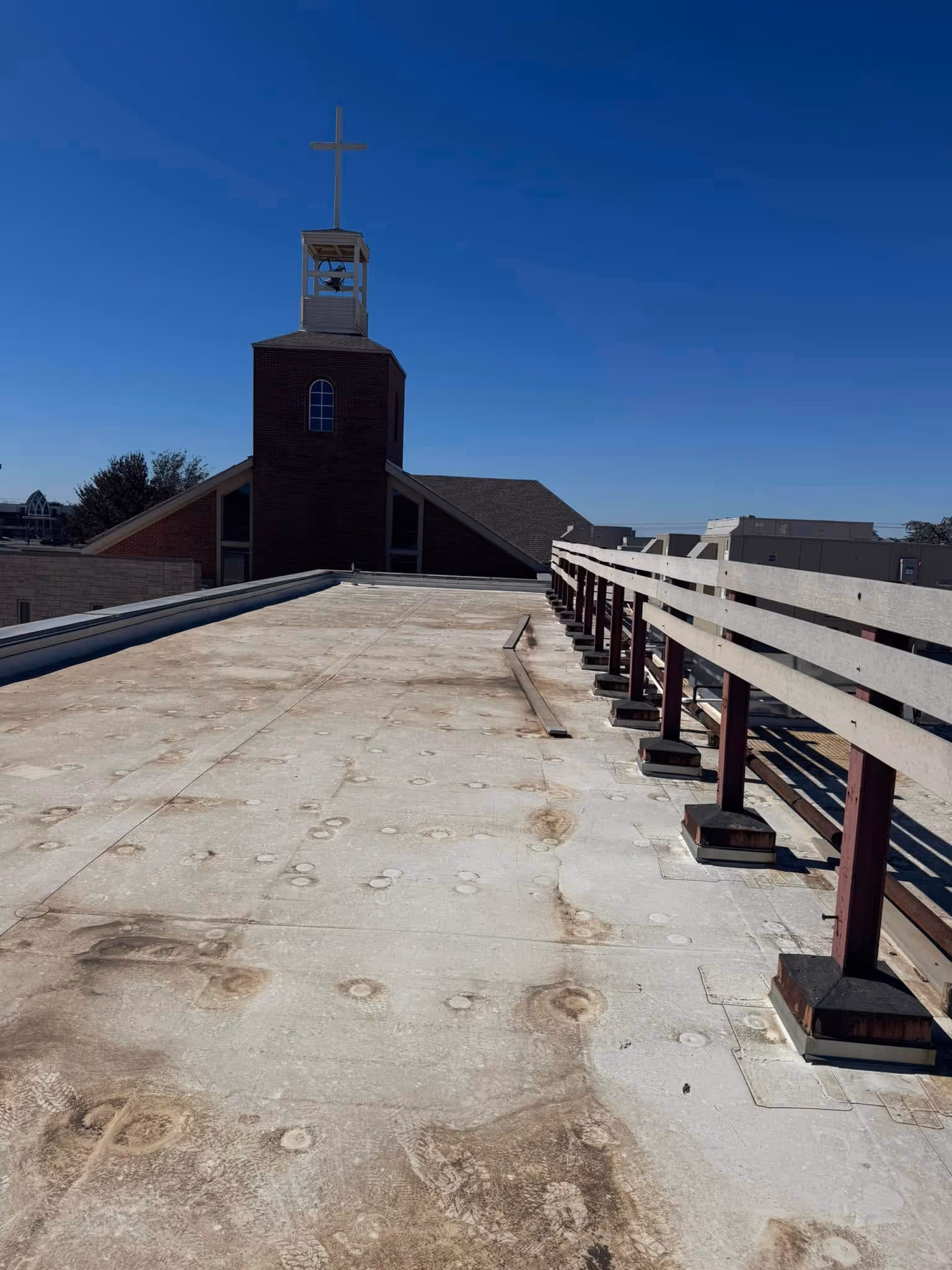 Flat rooftop with dirt marks and footprints, featuring a wooden railing on the right and a church steeple with a cross in the background under a clear blue sky.