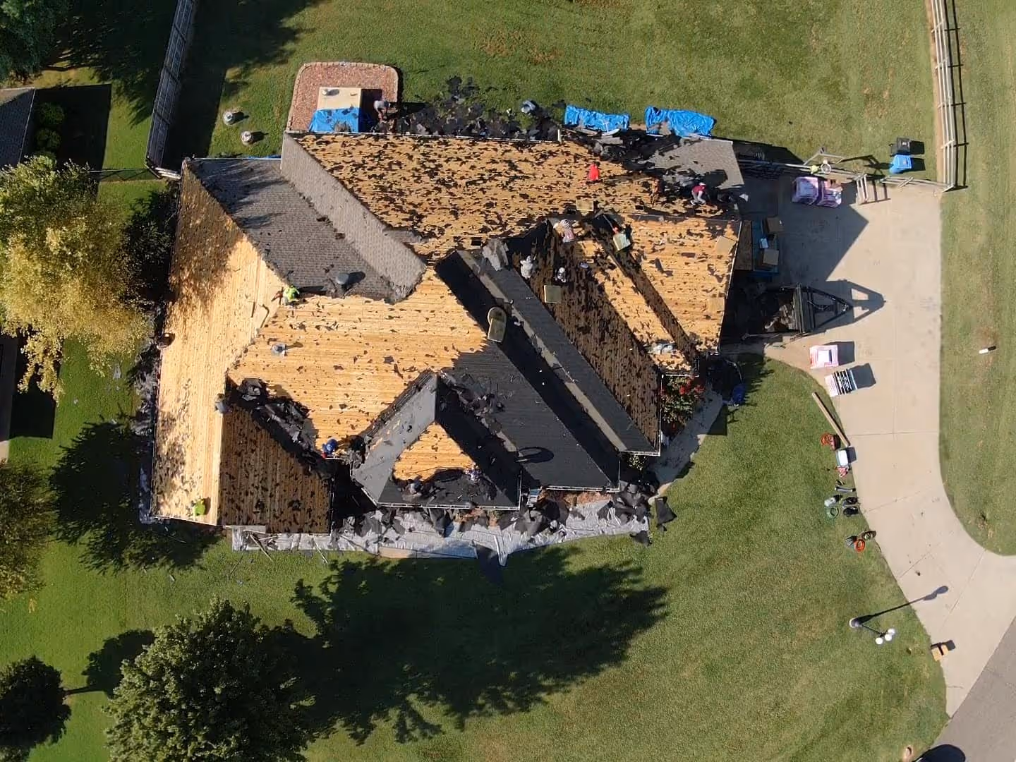 Roofing by Prodigy - Aerial view of a house roof undergoing repair with workers removing old shingles and debris on the wooden roof deck.