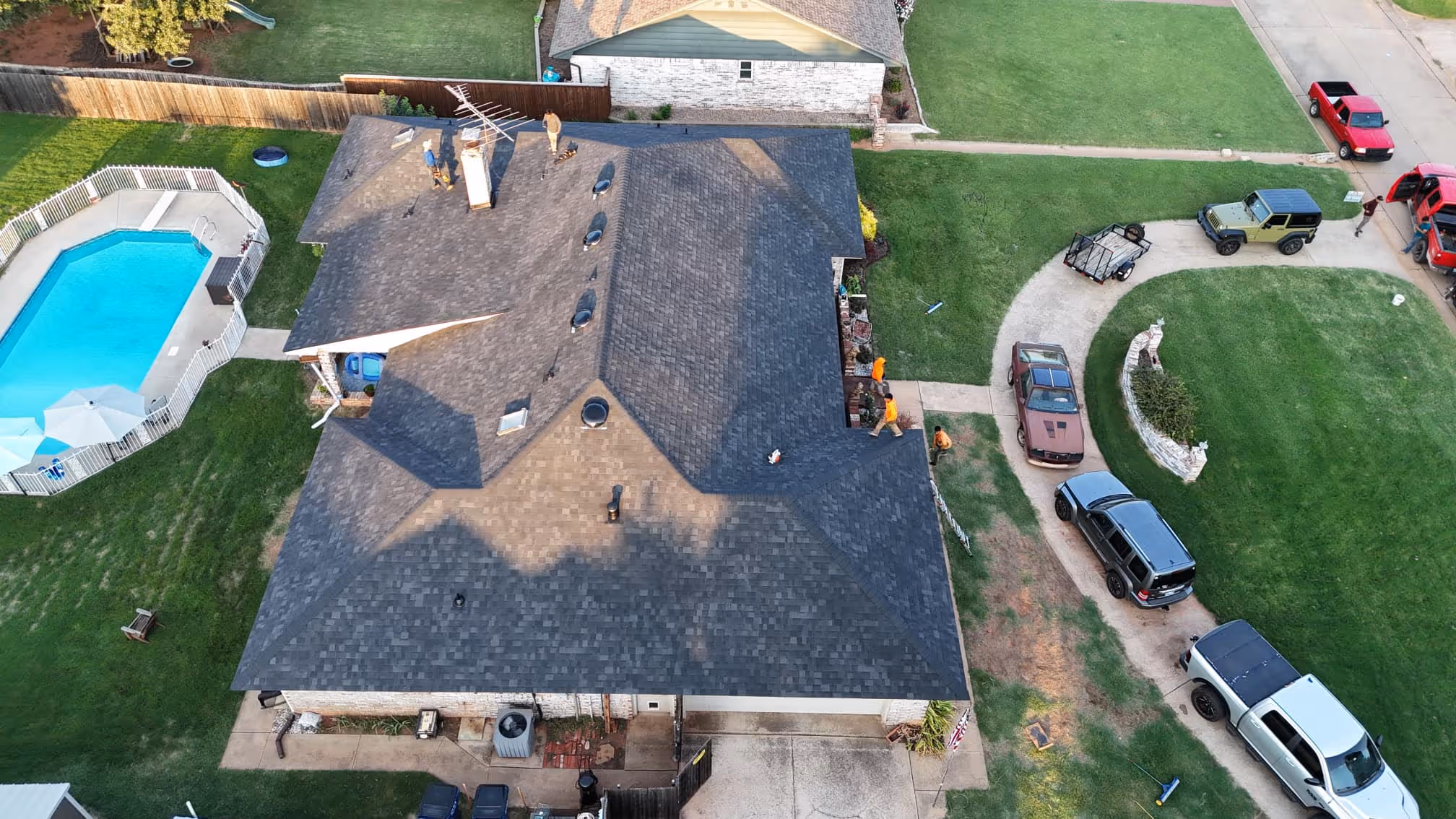 Aerial view of a large residential home with a dark gray roof, three workers on the roof, a fenced swimming pool, and several cars parked on a curved driveway.