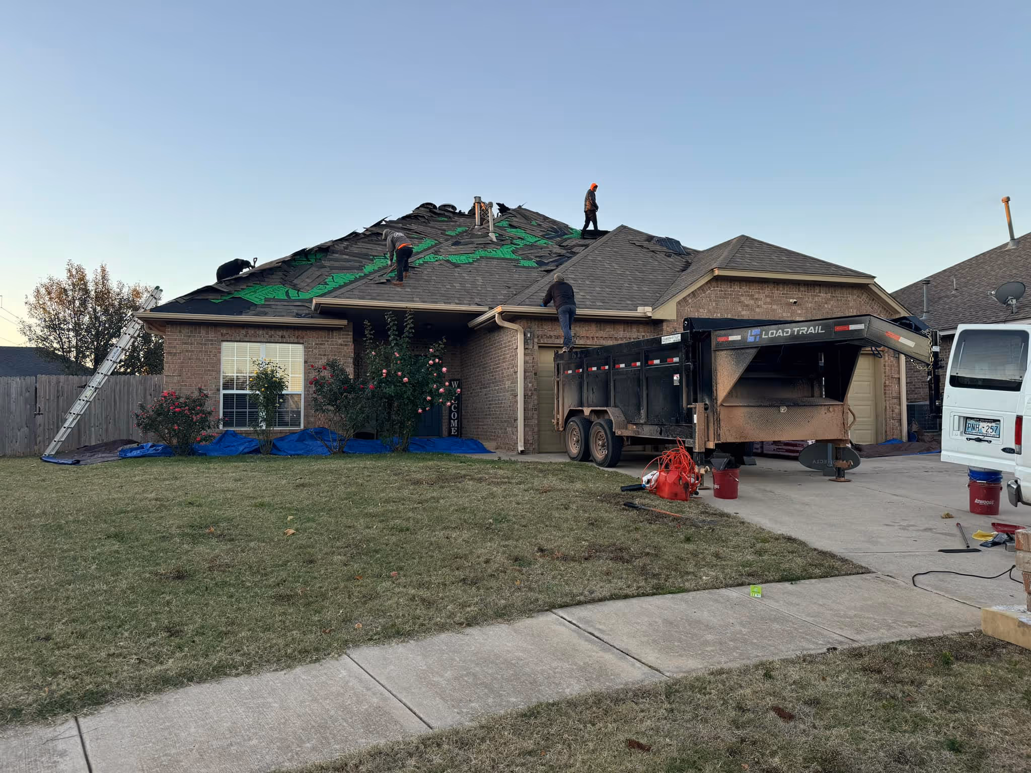 Workers repairing the roof of a brick house with roofing materials and a black Load Trail trailer parked in the driveway.