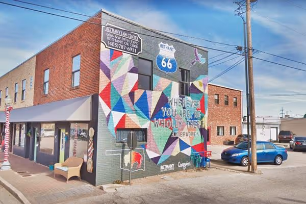 Colorful geometric mural on a brick building with Route 66 sign and text that reads 'Wherever you are, be wholly there' in Bethany, Oklahoma.