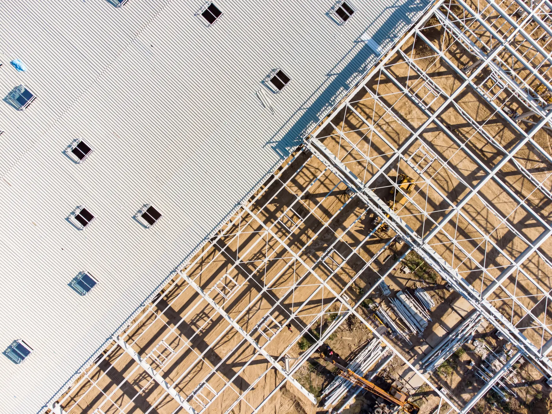 Aerial view of a warehouse construction site showing steel framework on the ground next to a partially completed corrugated metal roof with skylights.