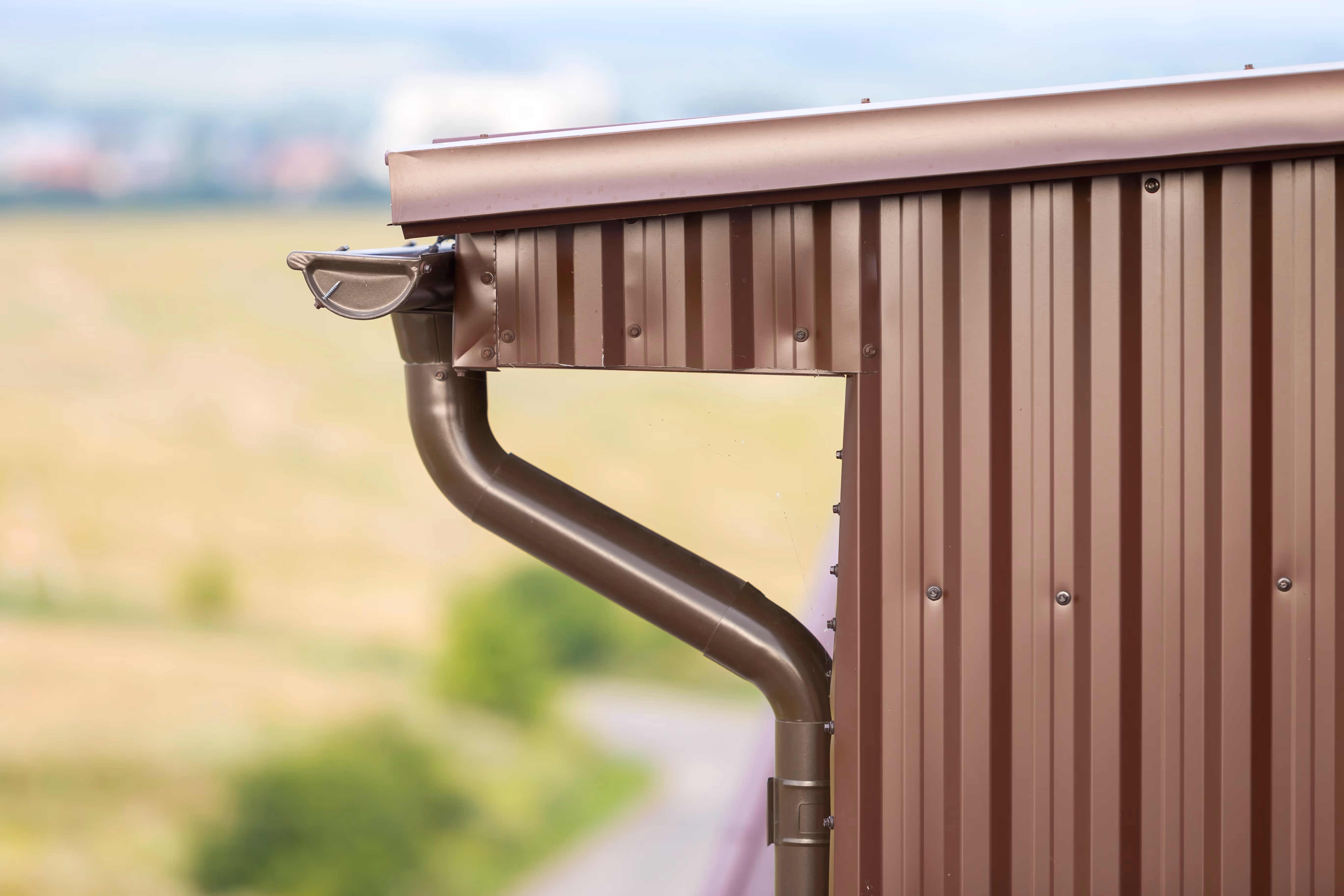 Close-up of a brown metal gutter and downspout attached to a corrugated metal building corner.