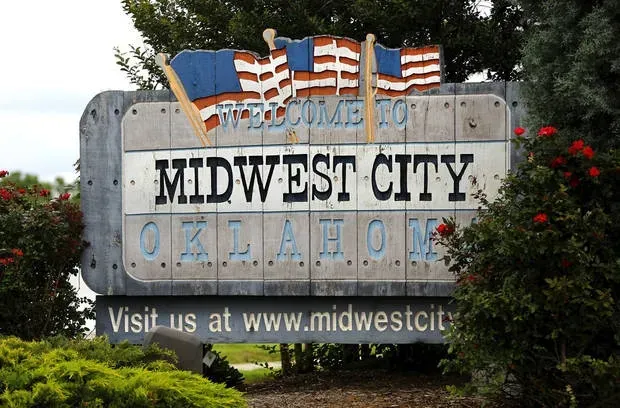 Stone "Welcome to Midwest City, Oklahoma" sign with American flags and red flowers.