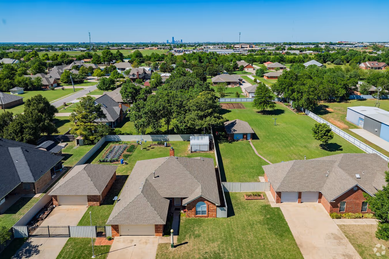 Aerial view of suburban houses with fenced yards, green lawns, trees, and a city skyline in the distance under a clear blue sky.