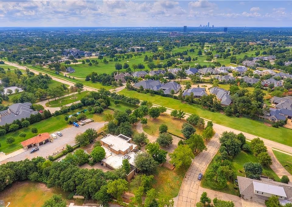 Aerial view of a green suburban neighborhood with houses, trees, roads, and a city skyline in the distance under a partly cloudy sky.