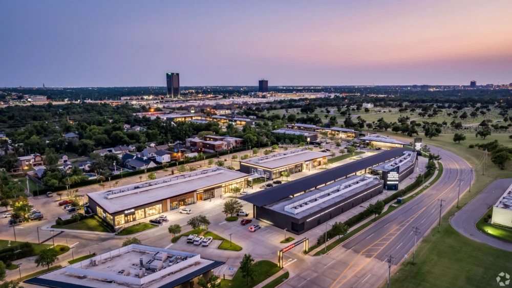 Aerial view of a modern shopping plaza with multiple buildings, parking lots, surrounding greenery, and a road at dusk.