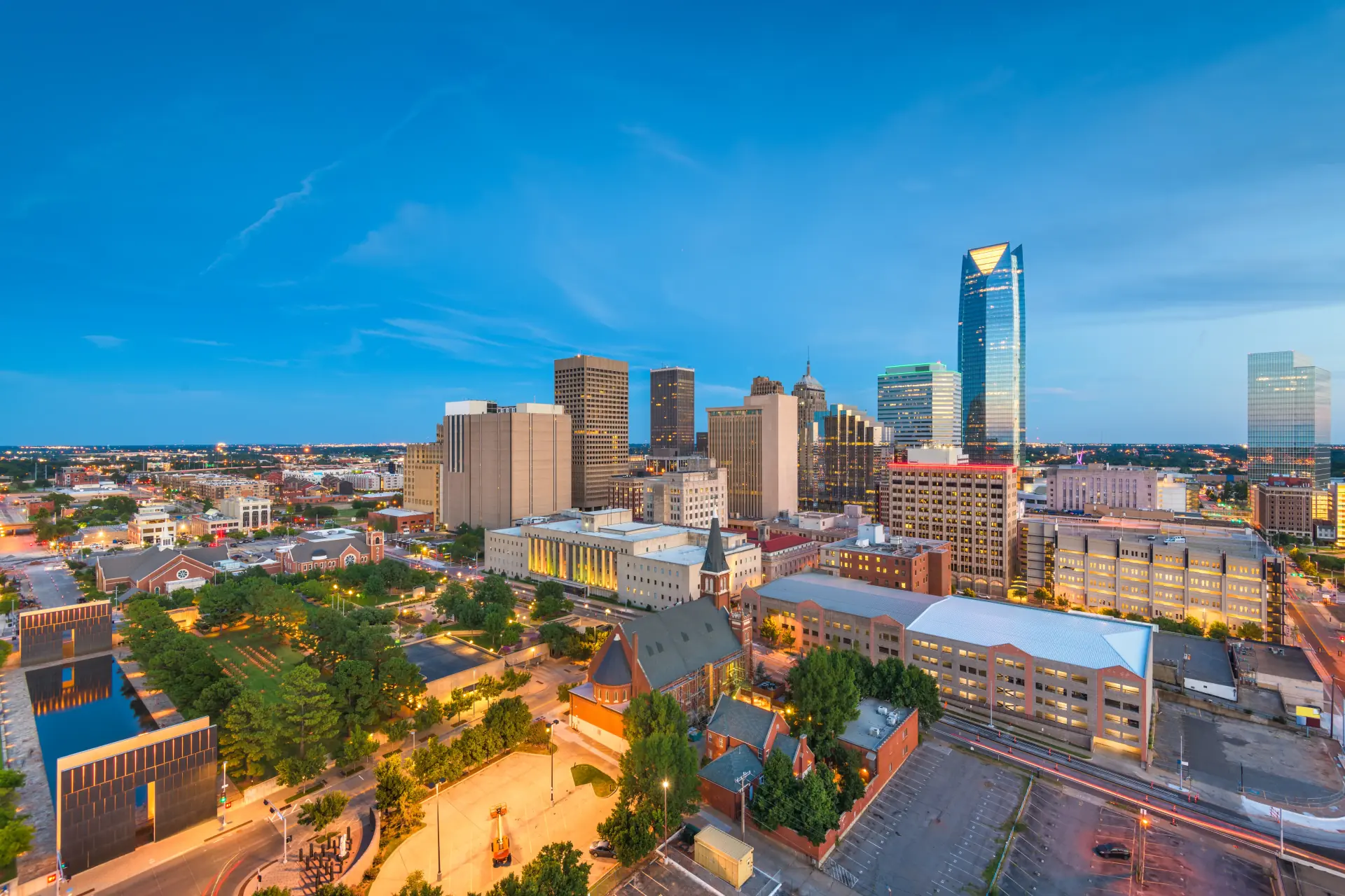 Roofing by Prodigy - Aerial view of downtown Oklahoma City skyline at dusk with illuminated skyscrapers, green park, and clear blue sky.