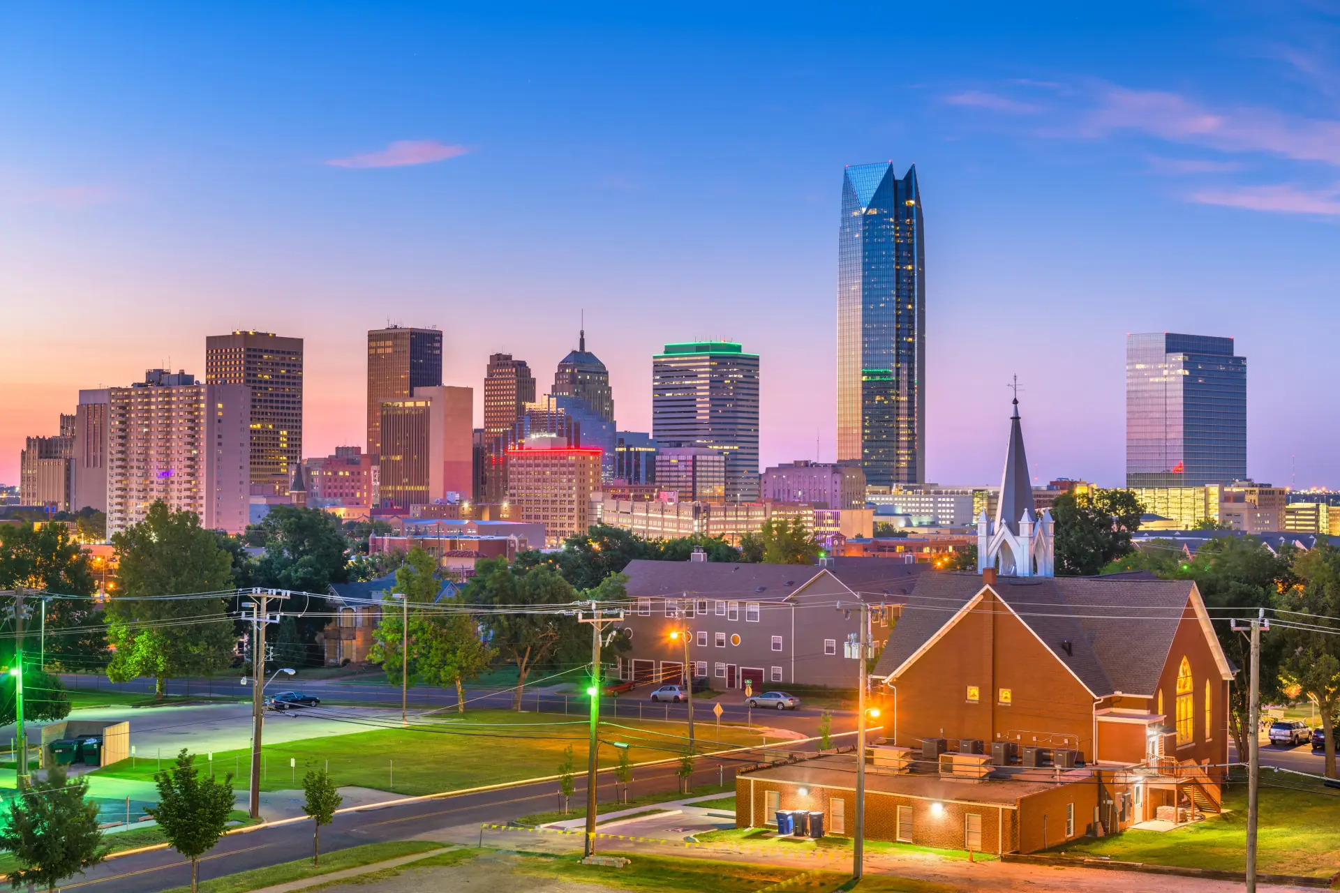 Oklahoma City skyline at dusk with illuminated skyscrapers, a church with steeple in the foreground, and a clear blue sky.