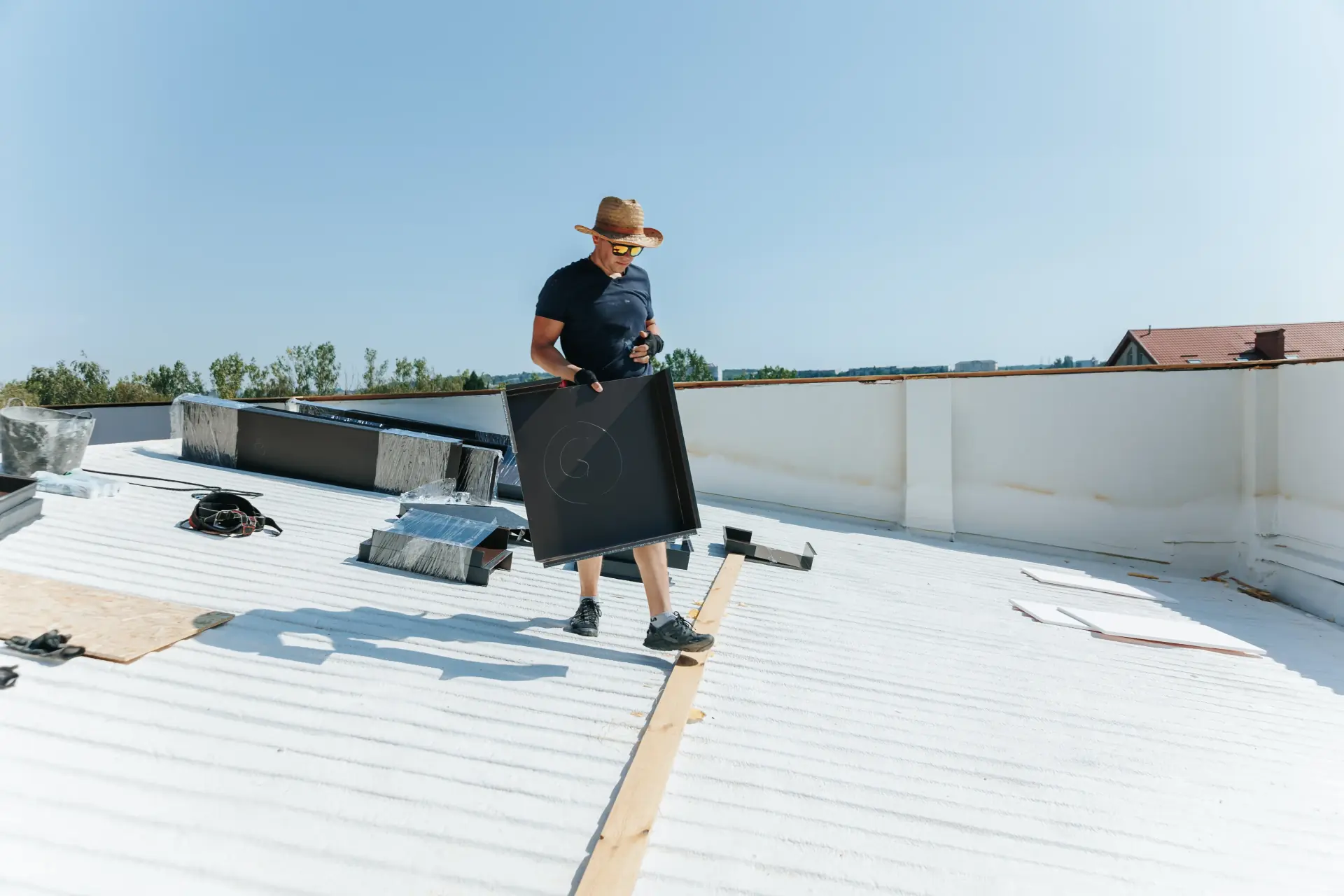 Construction worker wearing a straw hat and sunglasses carrying a metal sheet on a bright rooftop under clear blue sky.