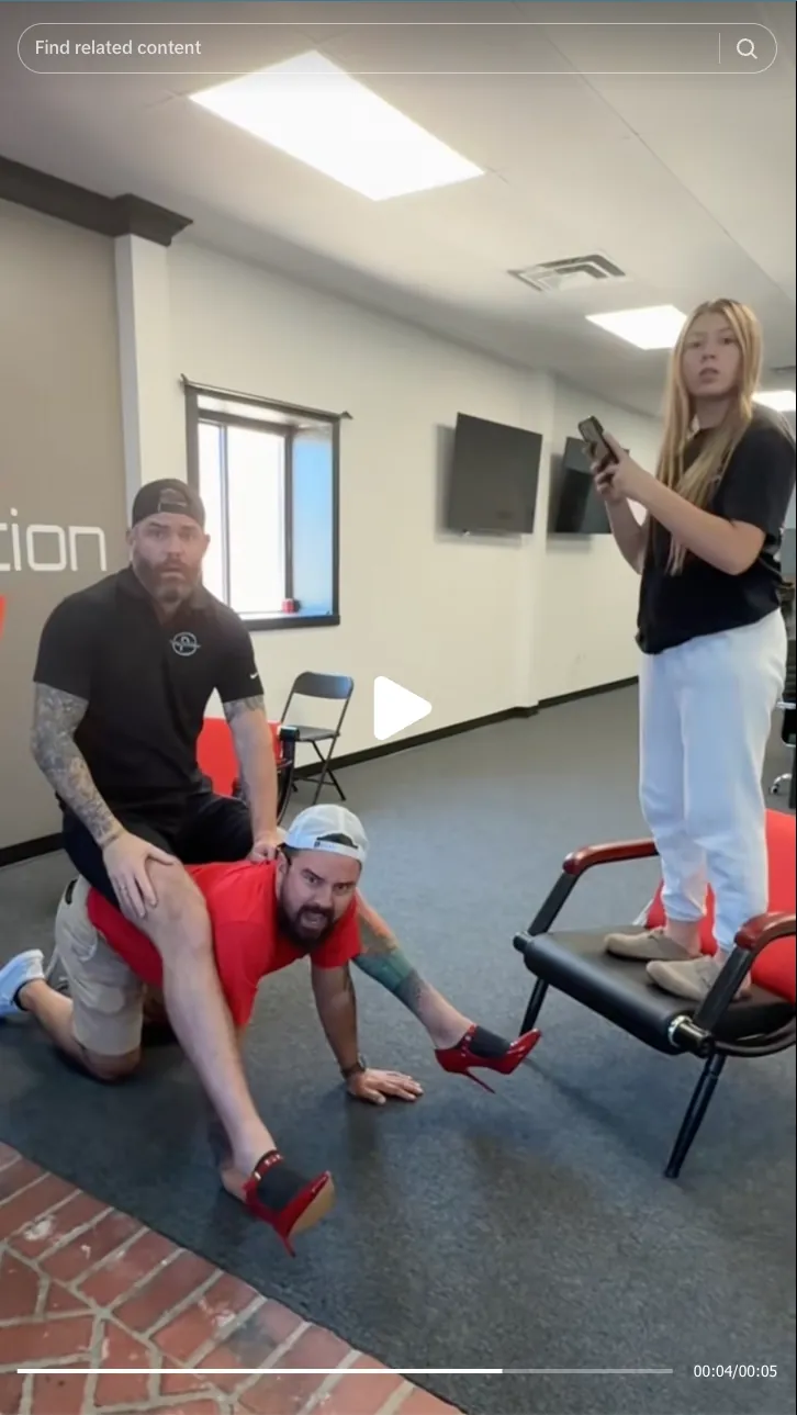 Roofing by Prodigy - Two men indoors, one kneeling with red high heels on, the other sitting on his back, while a woman stands on a chair holding a phone.